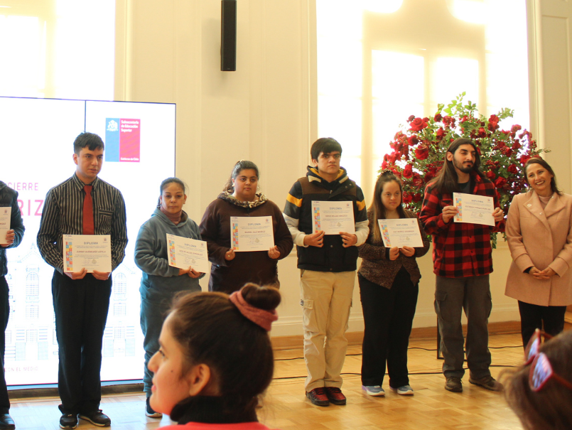 Beneficiarias y beneficiarios con sus diplomas, a un costado vicerrectora Jacqueline Páez. Sonríen para fotografía. 